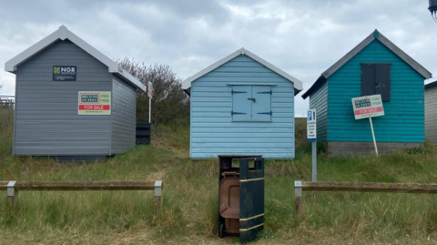 Three beach huts are in a line next to each other standing on grass. The one on the right is painted light grey and have a for sale sign on it. The middle one and the one on the right are painted blue and have a window. 