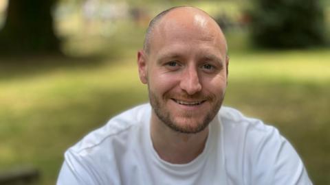 Saul Wardale smiling for the camera outdoors, wearing a white t-shirt.