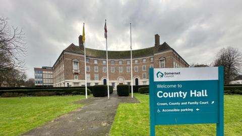 A large brown four-storey building with Georgian-style windows and three flag poles outside. There's a blue sign on the green grass at the front saying "Welcome to County Hall" and "Somerset Council".