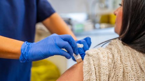 A woman sitting on a chair with dark hair. A healthcare worker in dark blue scrubs is holding up her sleeve and putting a plaster on her arm.