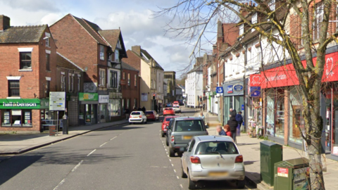 Shot looking down Nottingham Road in Alfreton with various shops visible on either side of the road, including Hall and Benson estate agents and a British Heart Foundation charity shop