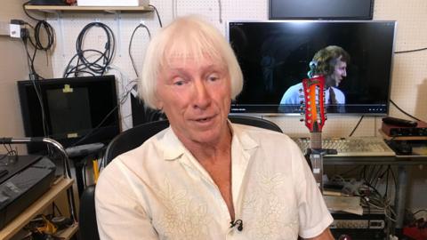 Bud Martin sat in a chair in a recording studio. He is wearing a white shirt and has white hair. Behind him is a guitar and computer equipment. There is also a TV.