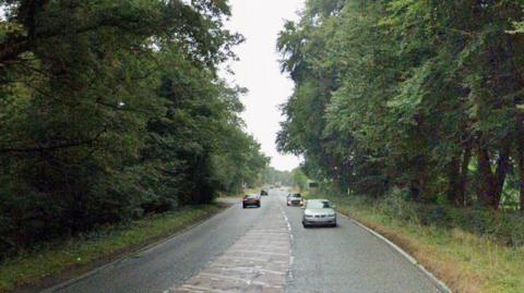 A Google street view image of a single carriageway main road in the countryside. Lots of trees are either side of the road and cars are travelling in both directions.
