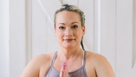 Dawn Blezard holds her palms together while wearing yoga clothes. She has blonde hair that has been french braided behind her head. 