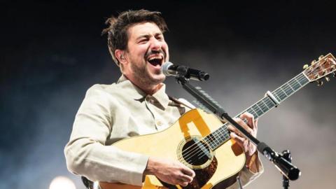 Marcus Mumford, wearing a light cream shirt, holding a guitar with his mouth open and a black and silver microphone in front of him