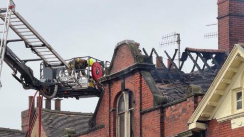 A firefighter working at the top of a building on an aerial platform. The roof supports of the building are charred and a chunk of the roof has fallen away. There are chimneys and aerials alongside the section of roof.
