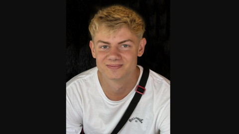 Fergus Lewis is smiling at the camera. He has blond hair and blue eyes and is wearing a white Billabong-branded T-shirt and has the strap of a bag across his shoulders. 