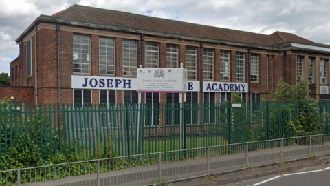 A view of Joseph Leckie Academy School. There is a redbrick building with a white sign on the walls reading Joseph Leckie Academy, although another sign on the grass cuts off the word Leckie from view. There is a green fence in front of the grass and the picture also shows a pavement.