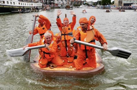 A group pf people in orange waterproof suits paddle their home-made raft towards the camera as part of the cardboard boat race at the Bristol Harbour Festival