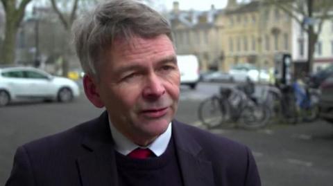 Andrew Gant being interviewed near St Giles boulevard in Oxford. There are bicycles and cars parked behind him.