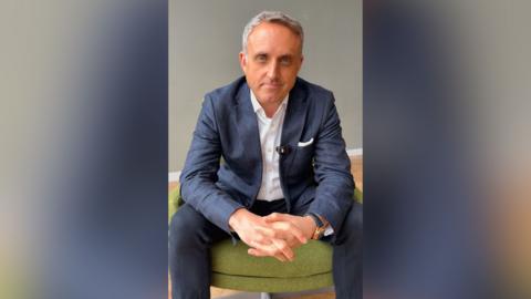 Alex Cole-Hamilton smiles at the camera as he sits on a green pouffe. He has silver hair, blue eyes and is wearing a white shirt and navy blue suit.