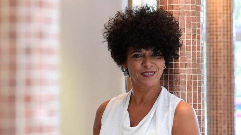 A woman with surly dark hair and a white top stares into the camera