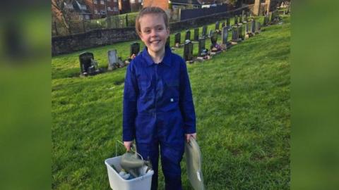 Ben standing in front of gravestones waering overalls and carrying a bucket of cleaning materials