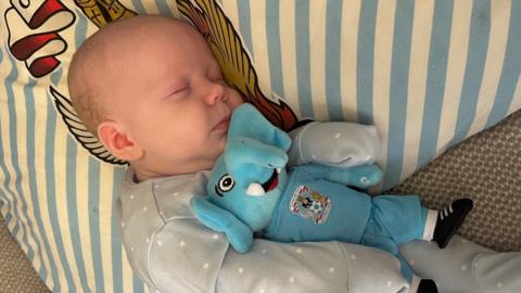 A newborn baby in a grey outfit with a toy elephant wearing a Coventry City kit