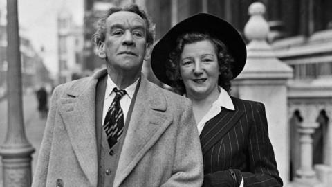 A black and white photo of a Arthur Lucan and Kitty McShane. They are middled-aged and handsomely dressed. Arthur Lucan has short hair and is wearing a blazer coat, cardigan, white shirt and striped tie. To the right, Kitty McShane, with shoulder-length curly brown hair is smiling with lipstick on, a wide-brimmed dark hat and is wearing a stripy, dark coloured blazer jacket and white unbuttoned shirt. A street can be seen behind them.
