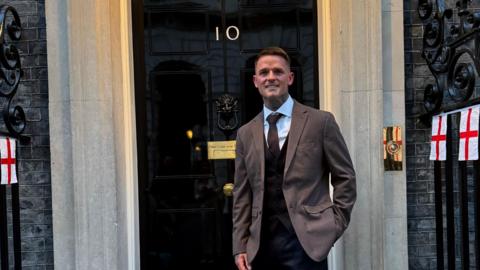 A smiling man stood outside the front door of Number 10 Downing Street. He is wearing a brown suit jacket with black waistcoat underneath, white shirt and brown tie. Attached to some black railings are St George's flags.