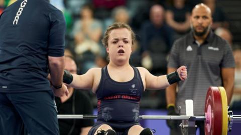 Charlotte McGuinness competing in a powerlifting event. She is seated on a bench, wearing a navy blue Great Britain competition uniform and wrist wraps, with arms extended outward as part of the setup for a lift. In front of her is a loaded barbell with red and yellow weight plates.