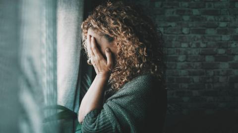 A woman with fair curly hair holds her face in her hands while standing next to a window