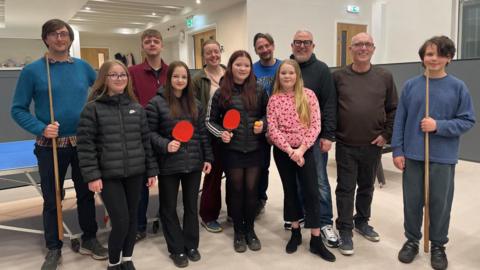 A group of young people pose together in a modern room with pool tables and a table tennis table. Some are holding cues while others hold bats. Older adults stand in a row behind them.