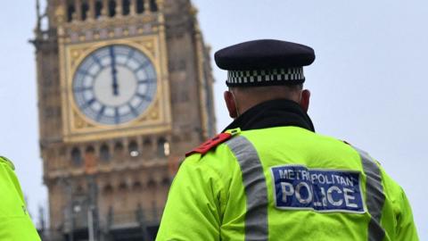 Close up view of the clock tower of the Palace of Westminster with a rear view of a police officer. He is wearing a high vis jacket and a cap.