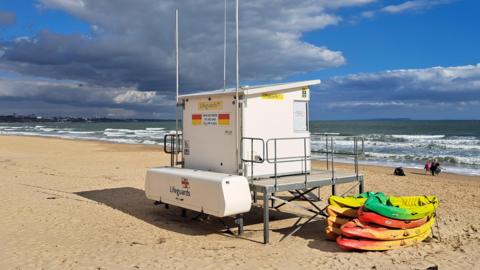 A closed lifeguard station sits on a beach looking out to sea. It's a nice sunny day but there are some large grey clouds in the sky. A stack of kayaks are next to the lifeguard station and there are a few people walking down by the sea