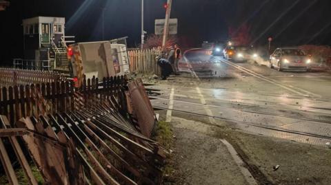 A damaged fence at a level crossing with a van on its side on rail tracks and a queue of traffic on a road.