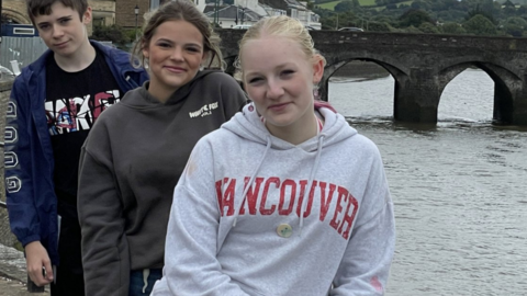 Three young people wearing blue, brown and grey hoodies with writing on then in front of a river and a bridge.