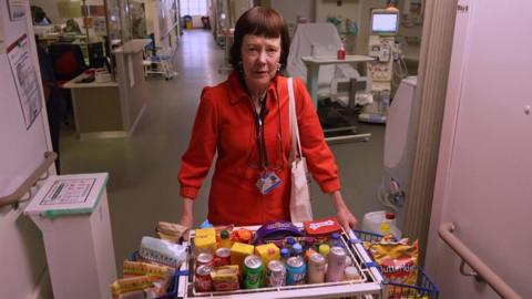 Honor standing in a hospital ward in a red coat. She has both hands placed on a trolley full of refreshments.