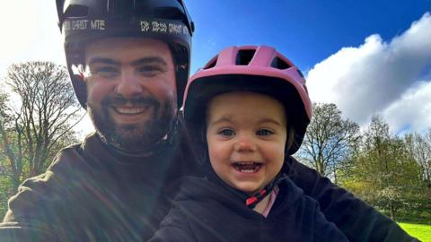 A man in his early thirties and little girl are smiling into the camera while sat on a bike wearing helmets. Green grass, trees and a blue sky with white clouds can be seen in the background. 