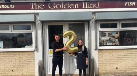 A man and a woman dressed in all black hold up a large golden balloon in the shape of the number two. They are standing in the doorway of a brick building. A sign above them reads "The Golden Hind".