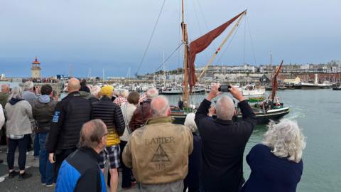 People standing by a harbour and watching a boat set off.