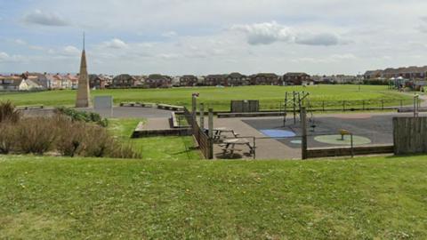 Google Streetview of Jubilee Gardens. There are large flat fields with houses in the distance, an obelisk to the left and a children's play area to the right.