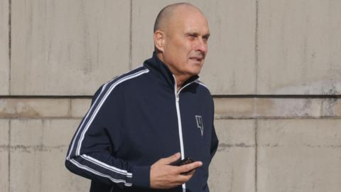 Derek Lammey, a man with a shaved head, pictured outside court at a previous hearing. He is wearing a navy sports jacket with white stripes on the sleeves and is holding a mobile phone.