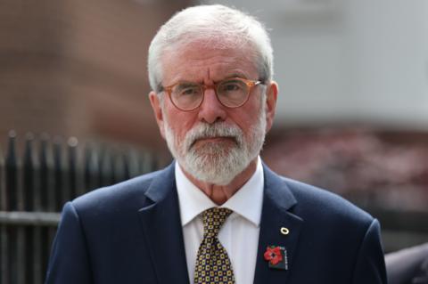 Gerry Adams is on a street. He has white hair and a beard. He's wearing orange glasses and a navy suit with a white shirt and a tie.
