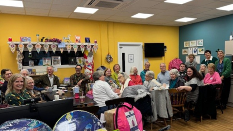 A long table with multiple people sat along it. They have turned to look at the camera and are smiling. The room has colourful walls decorated with crafts.