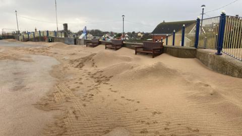 The photo shows a beach walkway. To the right hand side are piles of sand, which are almost as high as the wall in the background which is running adjacent to the path. There are three benches just in front of the wall. They are blocked off by the piles of sand. 
