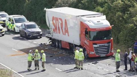 A red and white lorry marked with the logo of a transport firm called Sitra. The vehicle has been in a crash and is surrounded by debris and members of the emergency services