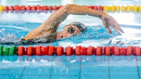 Image shows a young female swimmer partway through the front crawl with her right arm over her head, which is turned towards the camera. She is wearing dark goggles and a black swimming hat which contrast with the blue water - she is also swimming in a lane and the red lane dividers are either side of her.