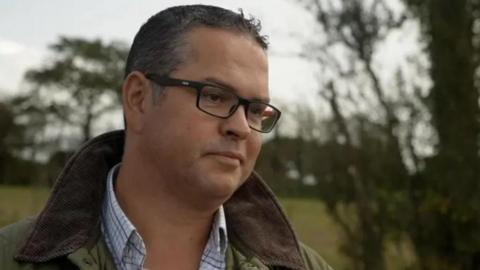 Marcus Calvani photographed from the shoulders up. Wearing a green coat, check shirt and glasses. He has short black hair and black framed glasses. A countryside scene is visible behind him. 