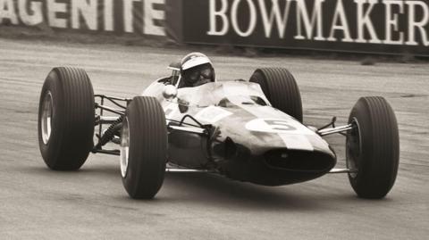 The Lotus 35 is shown in a black and white image on a race track with advertising boards behind it. The racing car is low to the ground with a slim-lined shape resembling a small jet aircraft with wheels smaller at the front than the back. A driver wearing an open faced helmet and goggles is in the cockpit.
