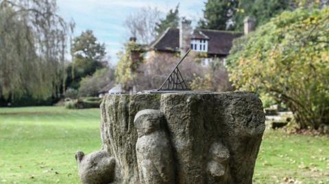 A weathered stone garden feature with a metal sundial set on top, engraved with numbers, stands in a lawn at Cotchford Farm, with shrubs, trees and the farmhouse visible in the background.