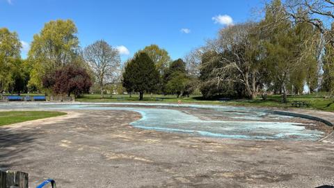 The drained paddling pool at Pickering Park it is grey concrete with a patch of blue in the middle and is surrounded by tall trees