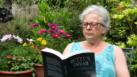A woman with white hair wearing glasses and a pale-blue patterned top reading a book named Mutti's Dream. There are plant pots and plants behind her.