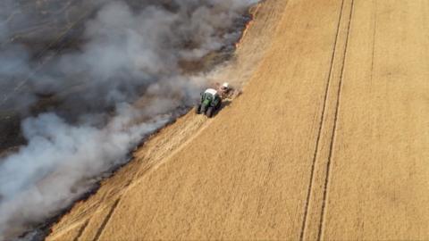 A drone shot of a tractor driving in a field separating a fire from untouched crops.