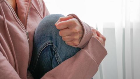 A woman, wearing a pink fleece and blue jeans, is sitting next to white curtains. Her head is not visible. Her posture is tense and she has a knee drawn up to her body.