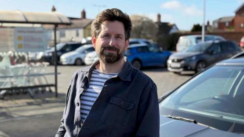 Gareth Wild stands in front of his car at the car park of his local supermarket. He looks at the camera and has dark brown hair and a beard. He is wearing a striped t-shirt underneath a navy blue shirt.