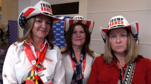 Three middle-aged women standing side by side wearing Trump Vance cowboy hats