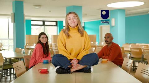 A woman wearing a yellow sweater is sitting on top of a meeting room table, smiling at the camera. Two other people are seated on chairs in the background.
