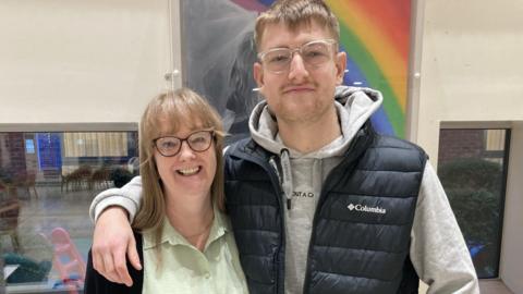 Will Postgate is wearing a gilet jacket over a grey hoodie and has his arm around Susan Baldwin who donated one of her kidneys to him. There is a piece of art showing a rainbow on the wall behind them.