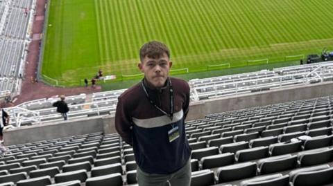 Pardoe Kendrick wearing a dark top, he is high up in the stands of a football stadium with the pitch in the background.